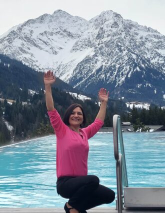 Heilerin Michèle Damonte mit hochgestreckten Armen vor einem Pool in einer Berglandschaft